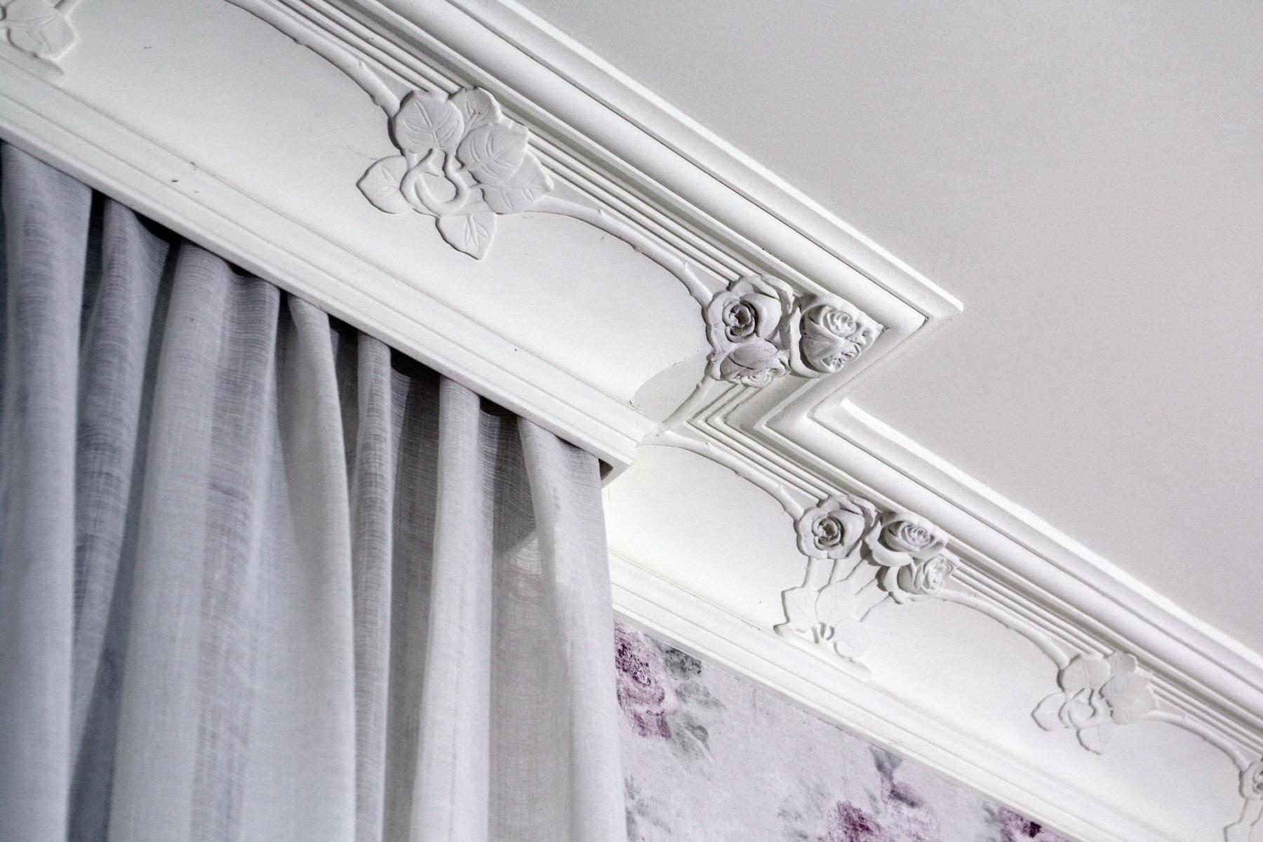 White ceiling cornice with sculpted rose motif corner over a sheer gray curtain and floral wallpaper